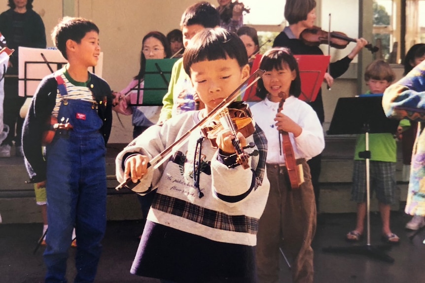 A young Brett playing violin with other children in the background.