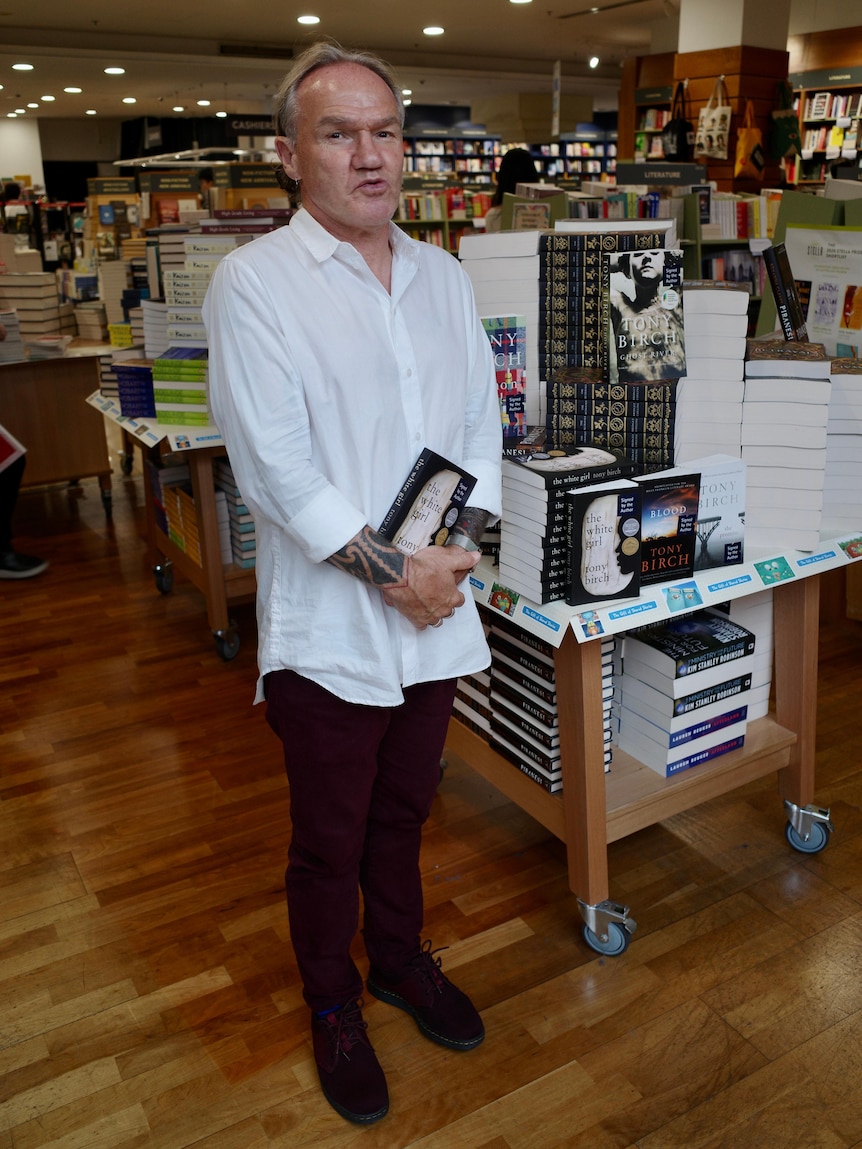 A man in a white shirt and black pants stands in a bookshop, holding a book.