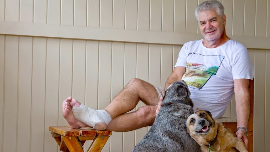 Wayne Shearman sitting with bandaged foot elevated, patting two cattle dogs.