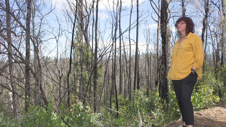 A dark-haired woman stands in front of burnt trees wearing a yellow shirt, looking into the distance.