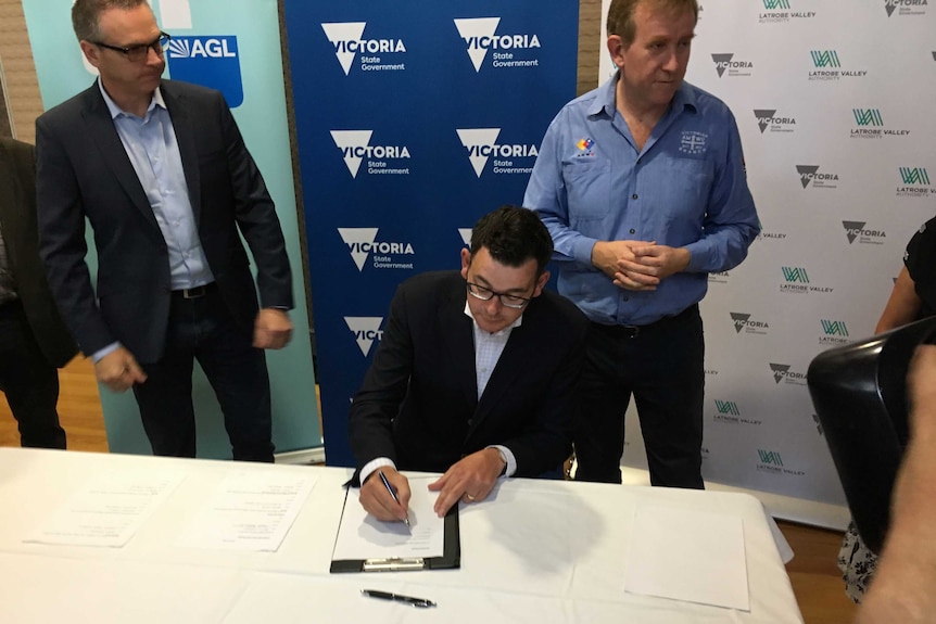The Victorian Premier Daniel Andrews is seated at a table and signing an agreement while two men look on.
