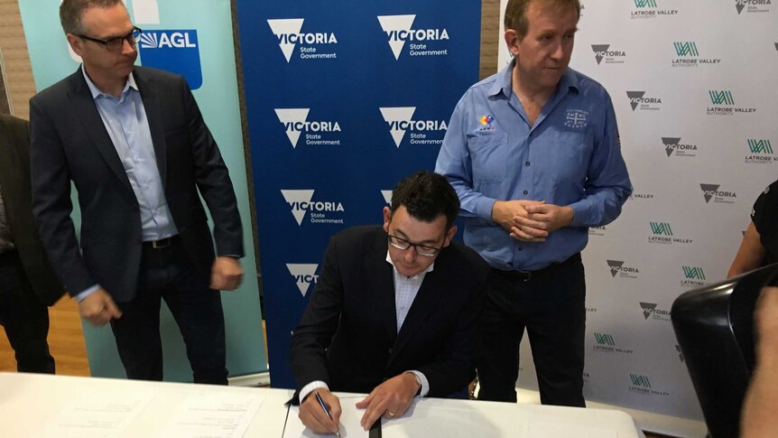 The Victorian Premier Daniel Andrews is seated at a table and signing an agreement while two men look on.