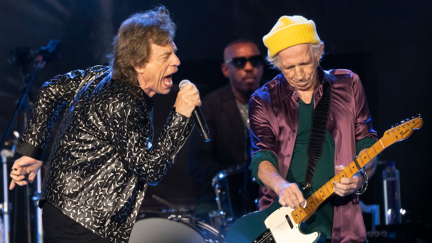 Rolling Stones lead singer Mick Jagger shouts into the microphone, with Keith Richards playing guitar and Steve Jordan on drums.