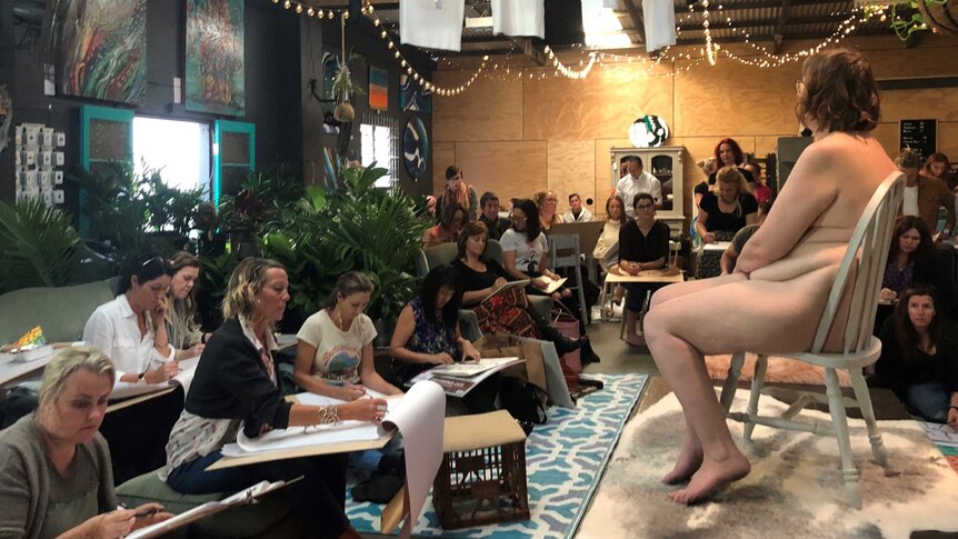 Alison Brinsley sits in a chair naked looking out over a crowd of artists