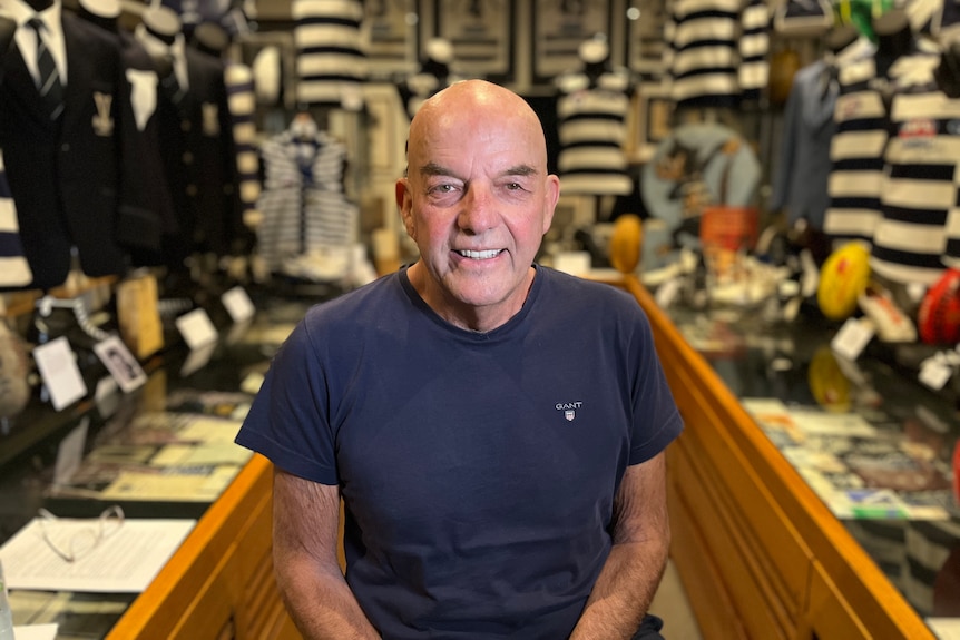 A man smiling at the camera surrounded by dozens of Geelong cats jumpers on the walls