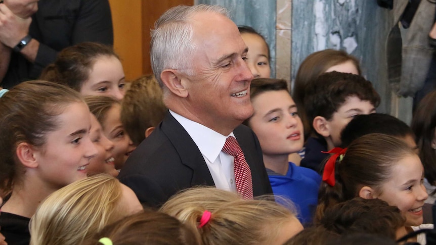 Malcolm Turnbull smiles cheerfully while wearing a black suit and a red and white tie sitting among primary school aged children