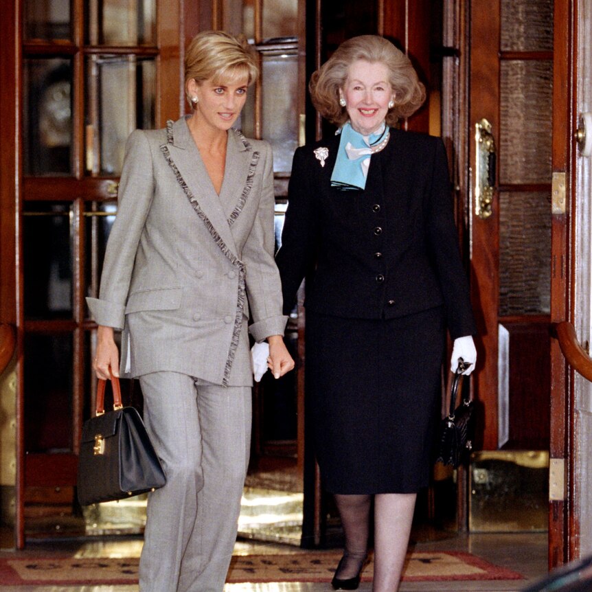 A blonde woman in a grey suit walks out of a building with an older woman in a black skirt suit 