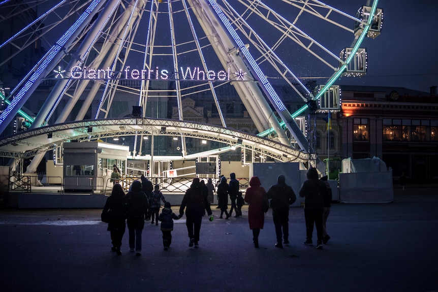 Families in silhousette walk towards a brightly lit ferris wheel at night as snow starts to fall in Kyiv