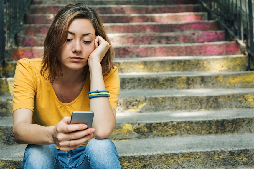 A girl sits on the stairs and looks sad as she looks at her phone 