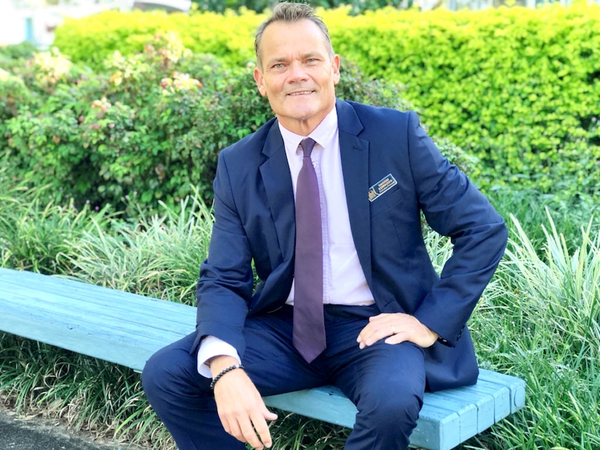 Man sitting on a bench, dressed in a suit, with greenery in the background and he's smiling gently at the camera.