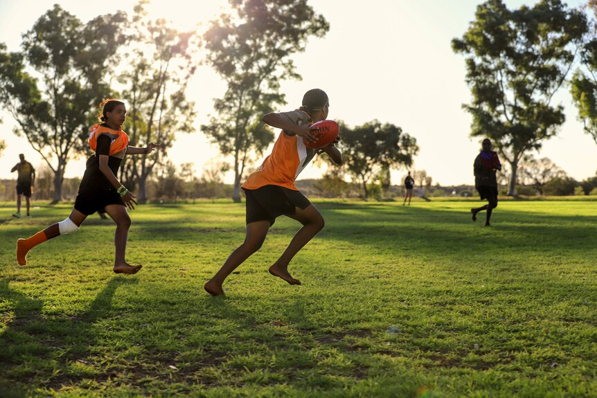 A young Aboriginal woman wearing a football uniform runs holding a ball, with a team mate running in support