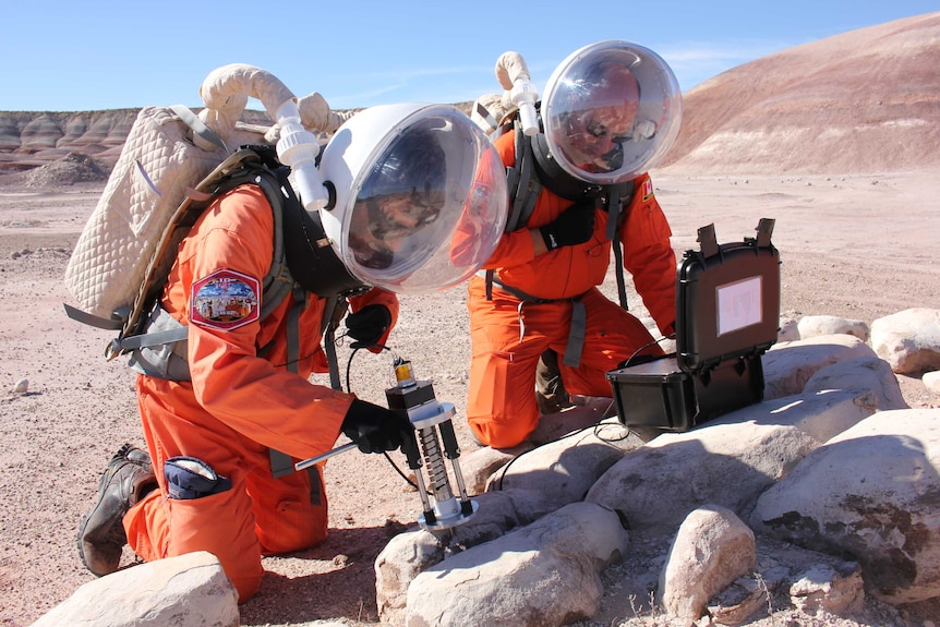 Tow men in space suits crouch by a rock