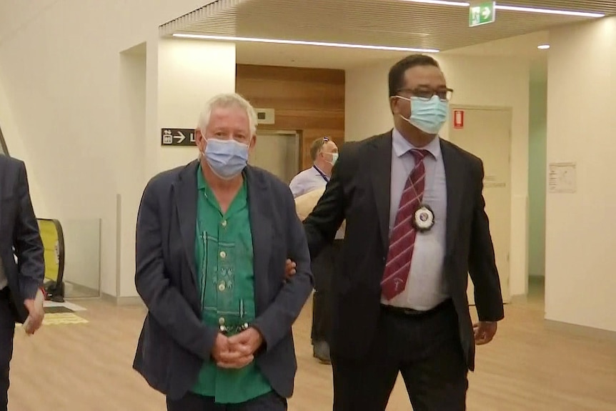 a man wearing a mask and handcuffs being escorted by two police through an airport