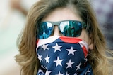 A woman with blonde hair and dark sunglasses wears an American flag over her mouth and nose.