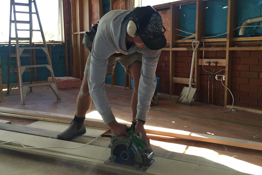 An apprentice builder leans over a piece of timber to cut it with a circular saw.