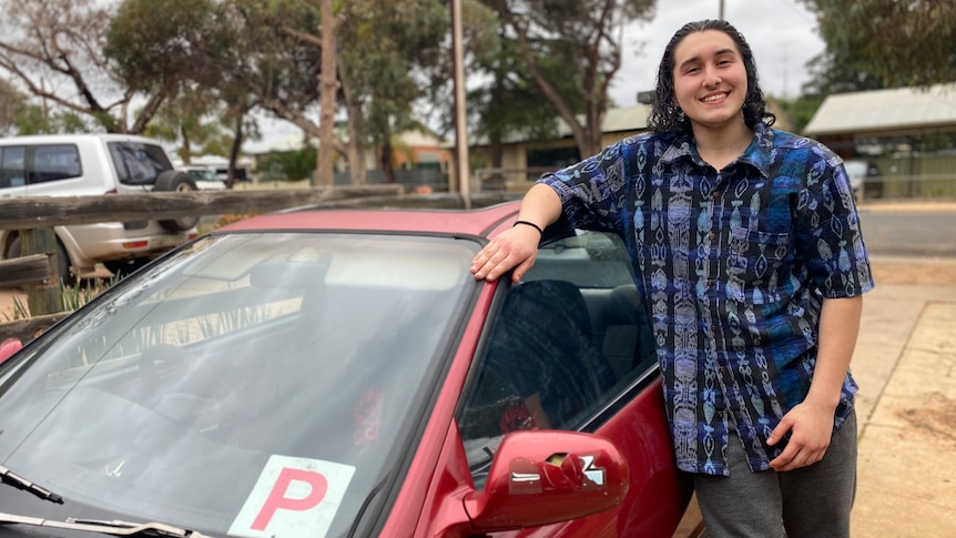 A young man has dark curly hair, he wears a purple and blue shirt. he smiles, hand resting on his red car with a red P plate 