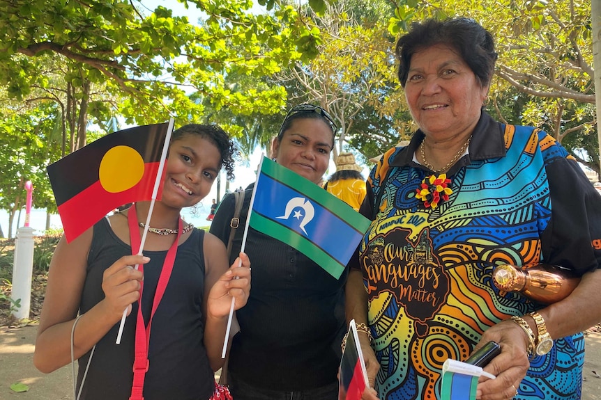 Three smiling women under green trees.  A young girl on the left holds an Aboriginal and Torres Strait Islander flag.