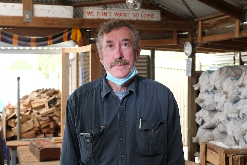 Man looking at camera, in centre with wood pile in background left and white bags of kindling stacked up on right
