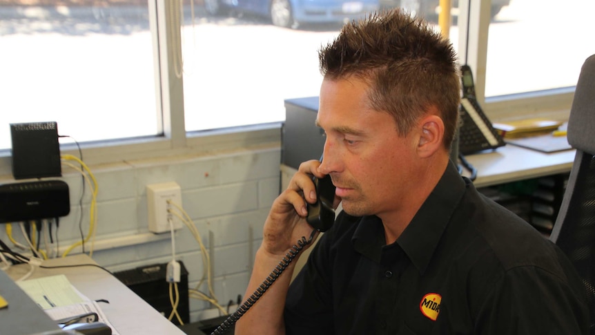 A man in a black workshop uniform uses the telephone at a desk in a mechanic's office.