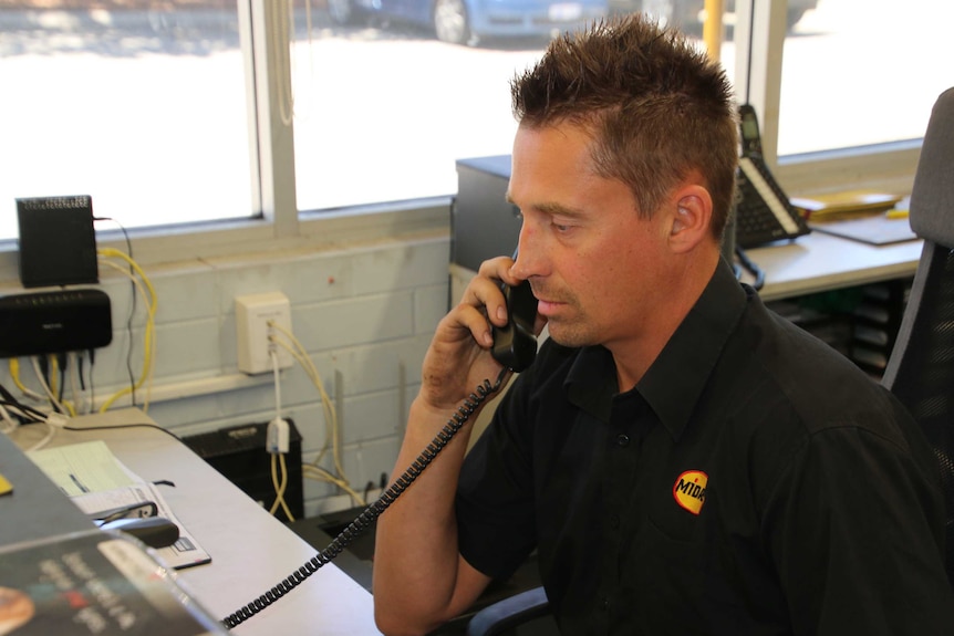 A man in a black workshop uniform uses the telephone at a desk in a mechanic's office.