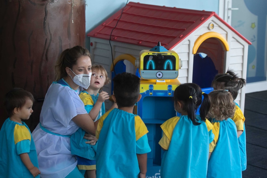 A woman in a face mask kneels down to show children in school uniforms how to use a robot thermometre.