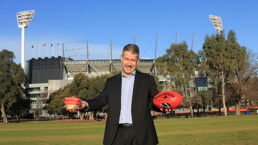 A man stands in a park outside the MCG, a large sports ground, holding an AFL football and three meat pies in plastic wrappers.