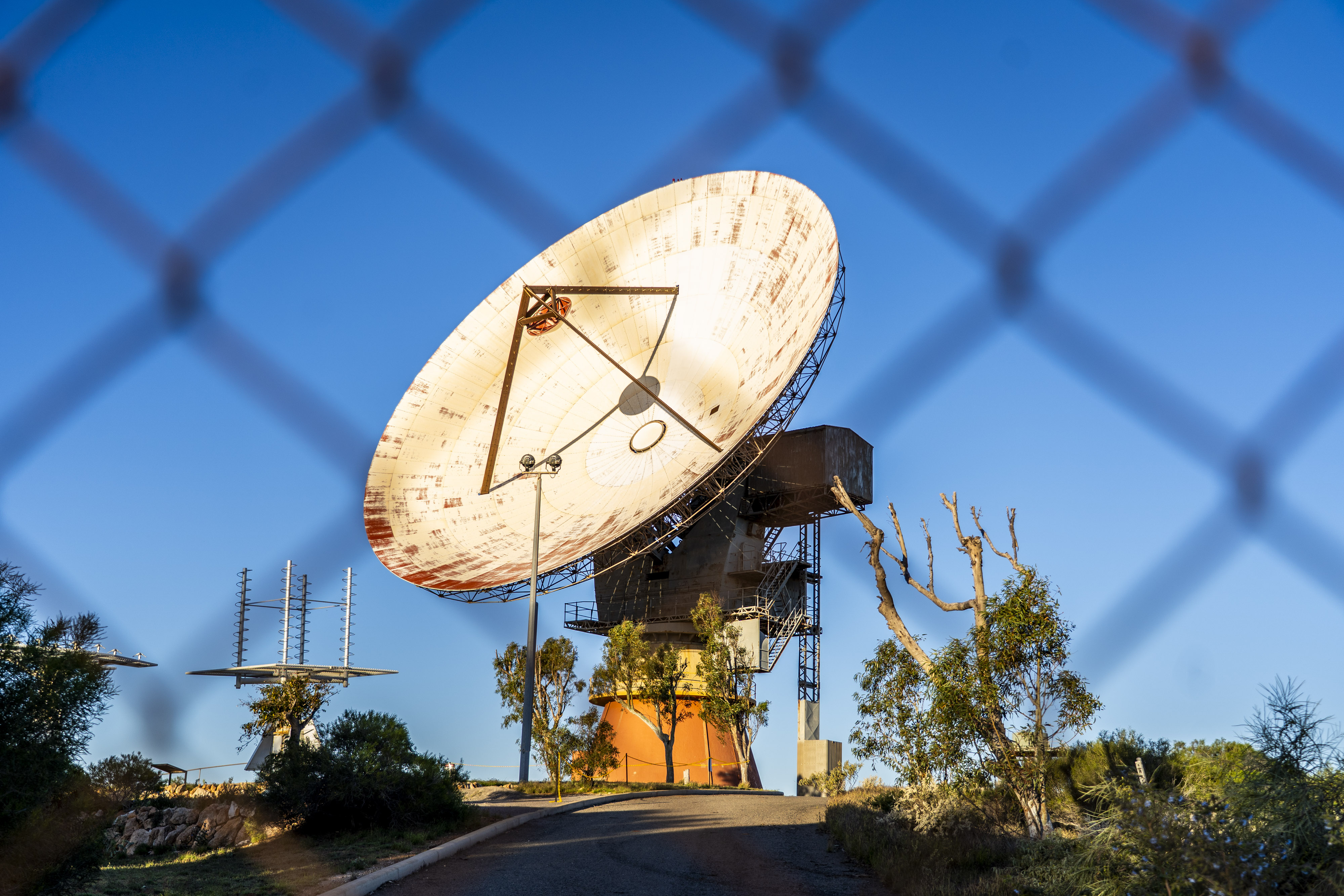 A huge weathered white painted satellite dish sits behind a fence on a hill
