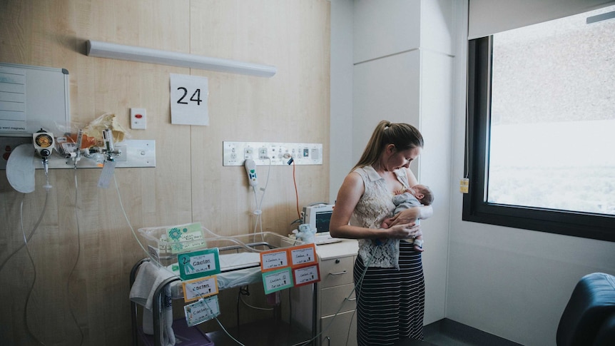 A young woman stands in a hospital ward with a tiny newborn baby in her arms.