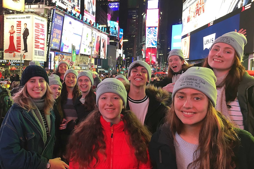 A group of teenagers in winter clothing with branded matching beanies, grin together amid the lights of Times Square.