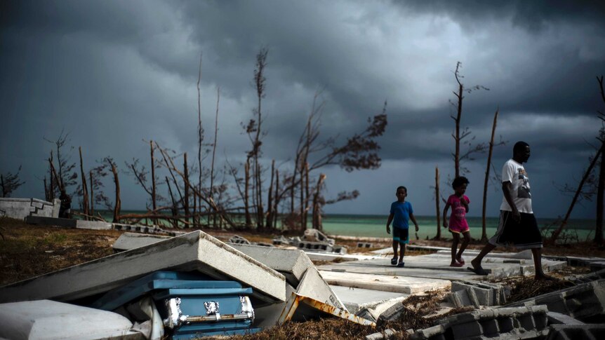 Residents on the island of Grand Bahama walk over the ruins of a building, with a shattered coffin in the front of the image.