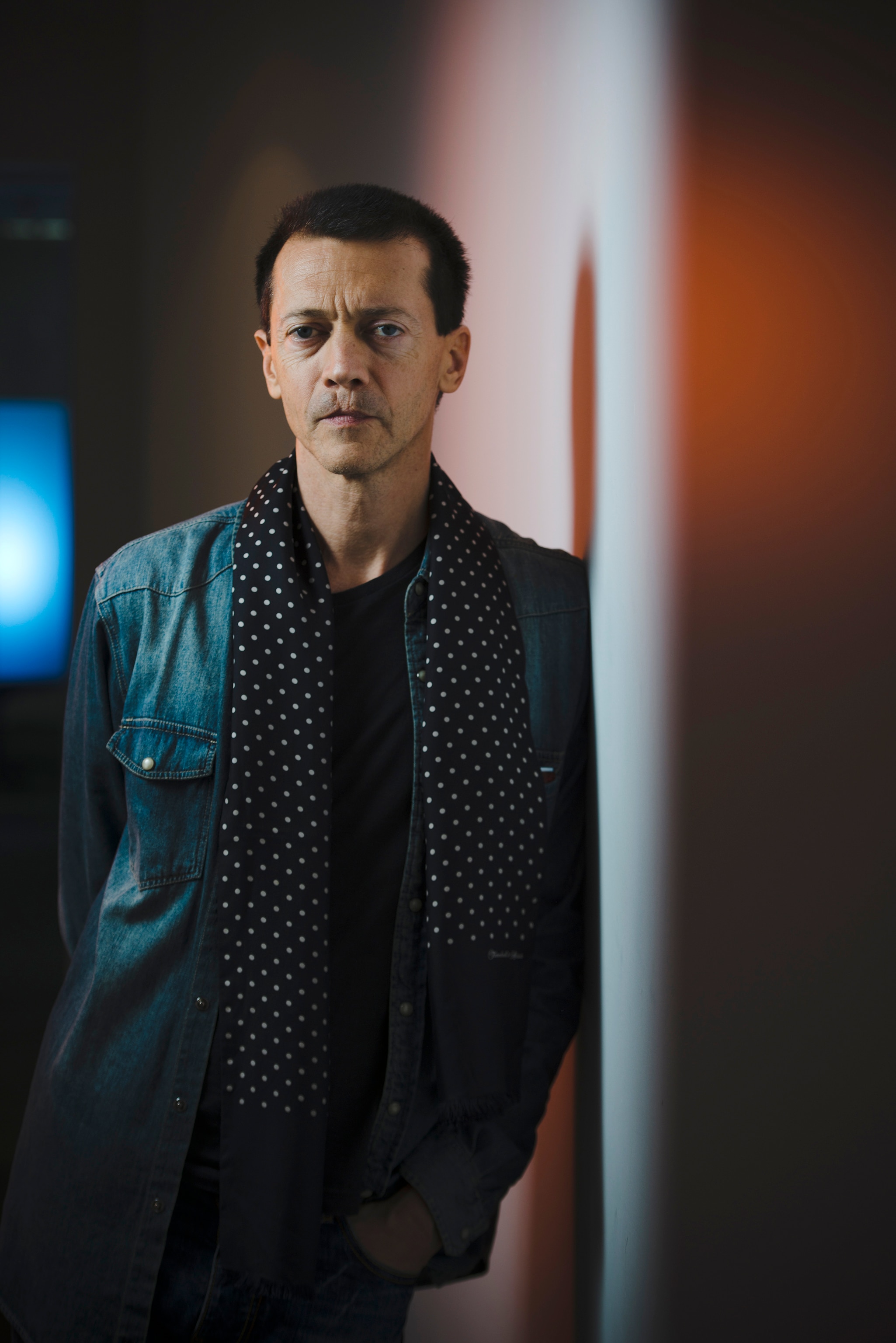 A middle-aged white man with short brown hair wears a denim jacket and a spotted black and white scarf leaning against a wall.
