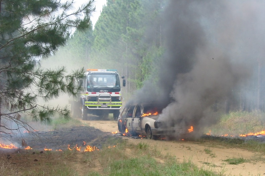 Car burning with a fire truck behind it, in the middle of a forestry plantation