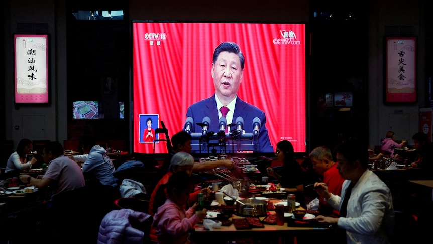 Xi speaking on a screen with red curtains in the background while people eat in a restaurant in the foreground.