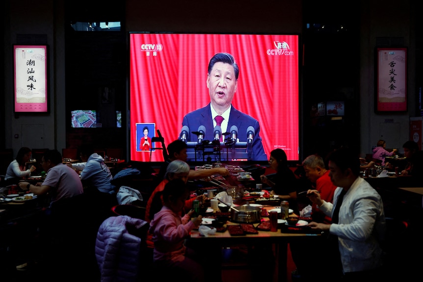 Xi speaking on a screen with red curtains in the background while people eat in a restaurant in the foreground.