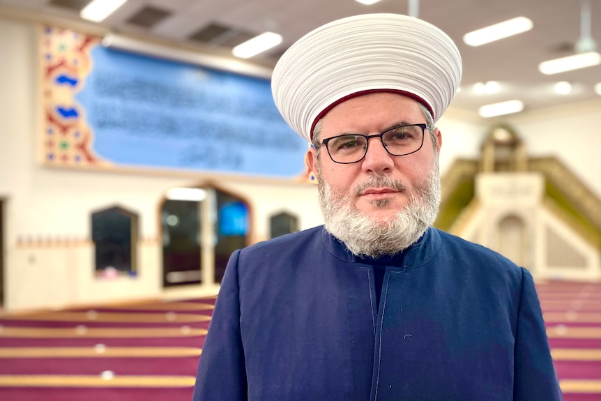 A man wearing a white clerical hat and blue coat looks towards the camera 