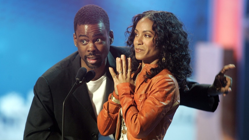 Chris Rock stands at a podium next to Jada Pinkett Smith, and speaks into a microphone while pointing to his left.