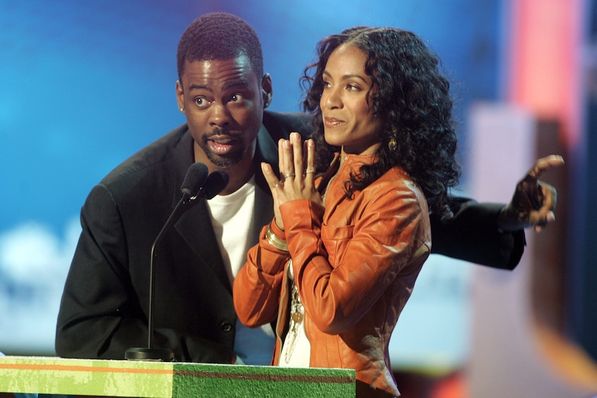 Chris Rock stands at a podium next to Jada Pinkett Smith, and speaks into a microphone while pointing to his left.