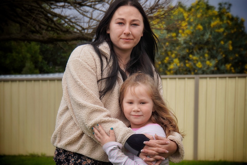 A mid-shot of a young woman with dark hair hugging her young daughter close and posing for a photo in a back yard.