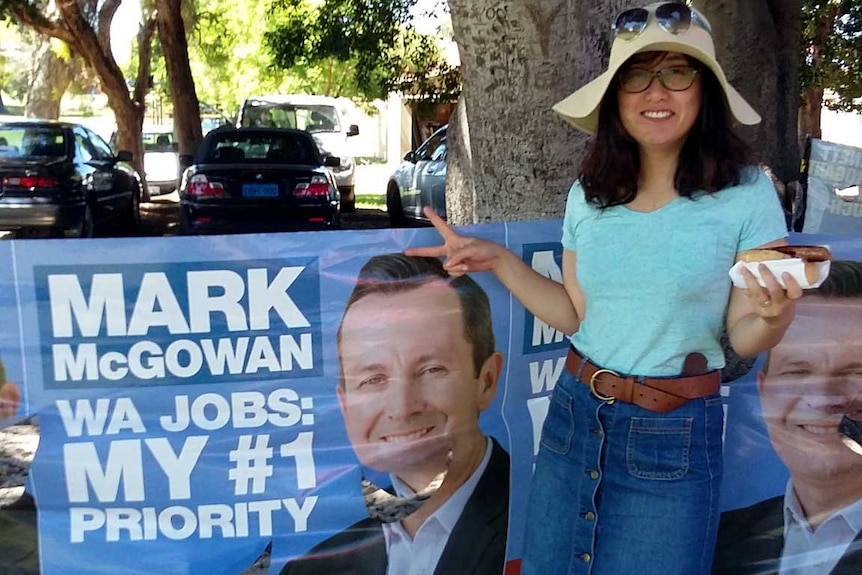 A woman in a hat carrying a hot dog poses next to an election banner featuring Mark McGowan.