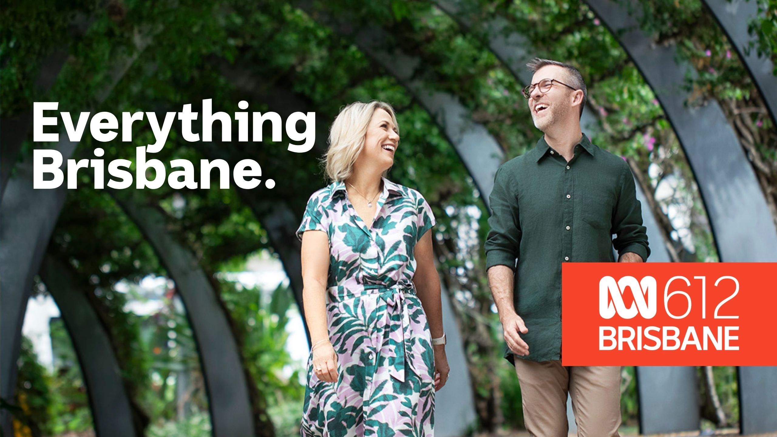 Two presenters walk through a leafy outdoor walkway with curved metal arches