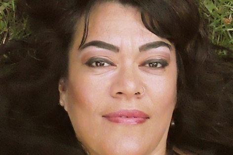 a Pasifika woman lies on the grass with her black hair around her and she is wearing a bright pink top