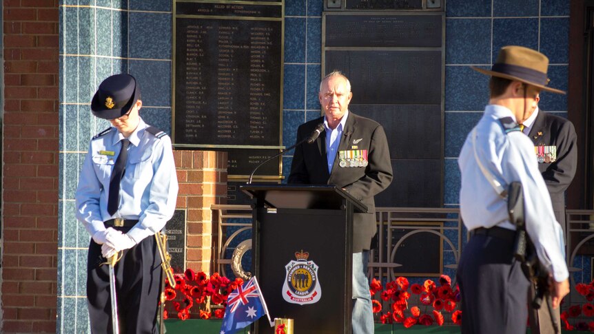 A veteran stands speaking at a podium, flanked by a guard of honour