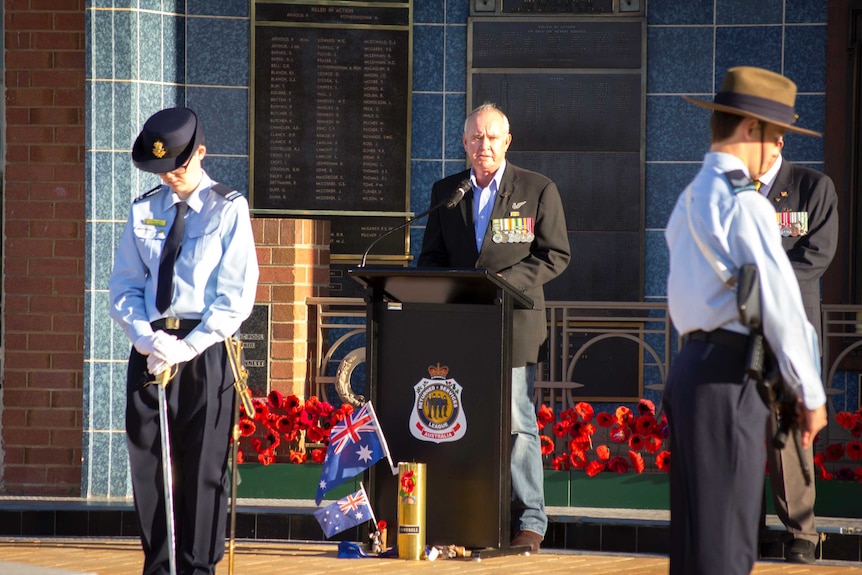 A veteran stands speaking at a podium, flanked by a guard of honour