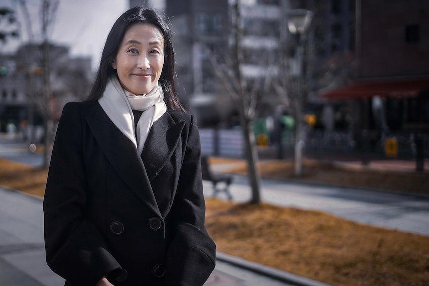 A Korean woman in a black coat and white scarf stands on a street in Seoul