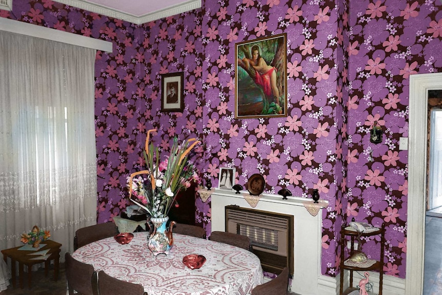 You view a dining room with bold purple and pink wallpaper with a flower motif framing 1970s-era furniture.