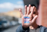 Closeup of the palm of the hand of a young person with a transgender flag painted in it, in front of their face