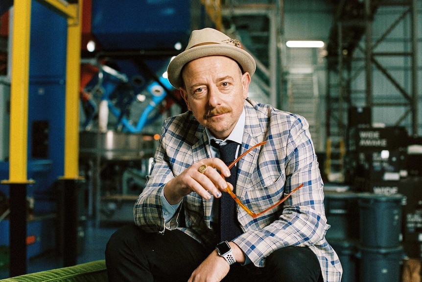 A man wearing a plaid jacket and hat poses for a portrait in a coffee warehouse
