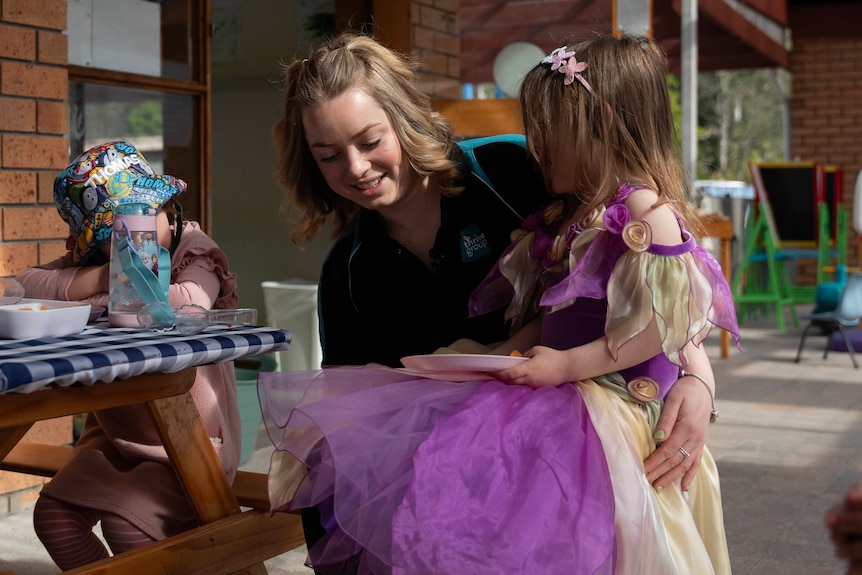 A child dressed in a princess dress is sitting on the lap of a childcare worker as they eat their morning tea.