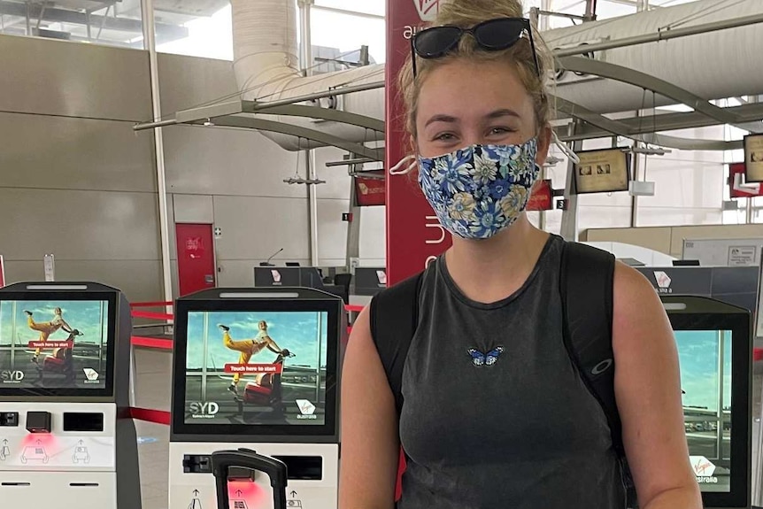 A young woman in a mask at the airport grins, blonde hair and bags around her