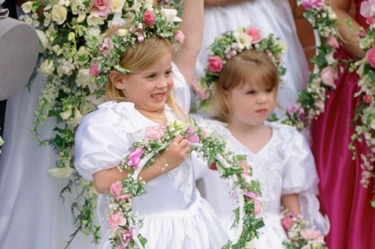 Two girls with blonde hair and flowers in their hair wear white dresses and hold a wreath of flowers.